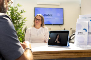 A woman wearing glasses and a white top sits behind a counter at a bank. There is digital signage behind her and brochures on the counter advertising "i-Bank". She is having a conversation with a Deaf man using Video Remote Interpreting (VRI) On-Demand via a tablet on the counter.