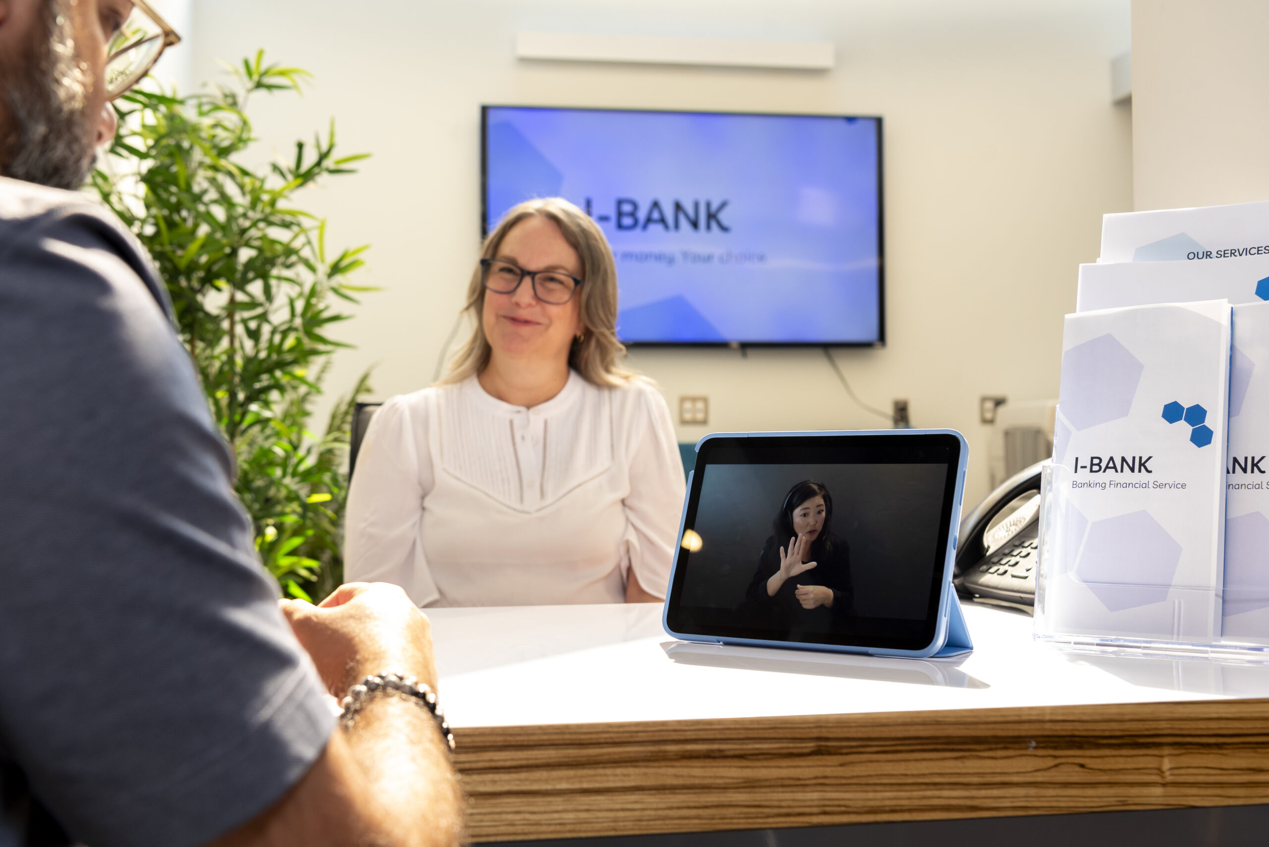 A woman wearing glasses and a white top sits behind a counter at a bank. There is digital signage behind her and brochures on the counter advertising 