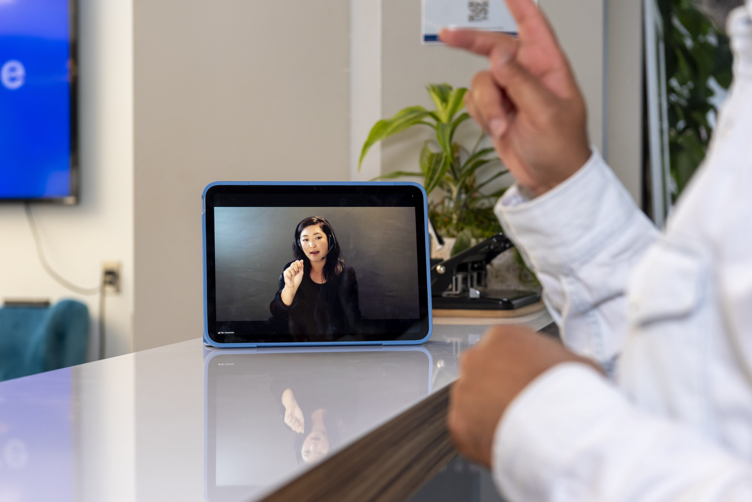 A tablet is sitting on a service counter showing a sign language interpreter. A man's hands and forearms are visible as he signs to someone off-screen using Video Remote Interpreting (VRI) On-Demand.