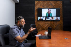 A man with gray hair and glasses signs with his team using Video Remote Interpreting (VRI) during a virtual meeting in a conference room. His laptop screen shows multiple participants, including an interpreter in a green blazer leading the discussion. A wooden panel wall with a large monitor forms the backdrop.