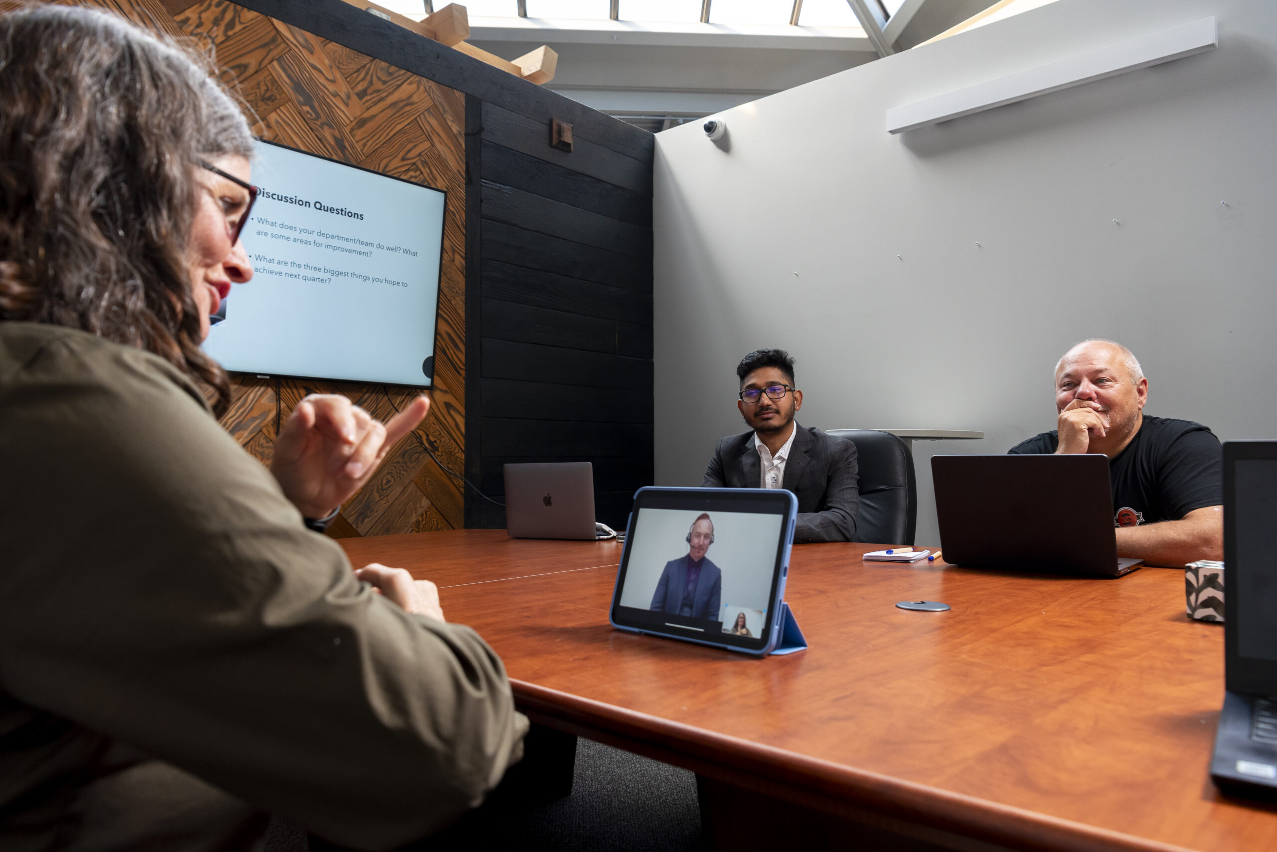 A woman in an olive green jacket signs in a workplace meeting while two colleagues, one in a suit and the other in a t-shirt, watch attentively. A tablet on the table displays a Video Remote Interpreter facilitating communication, and a large monitor in the background shows discussion questions.