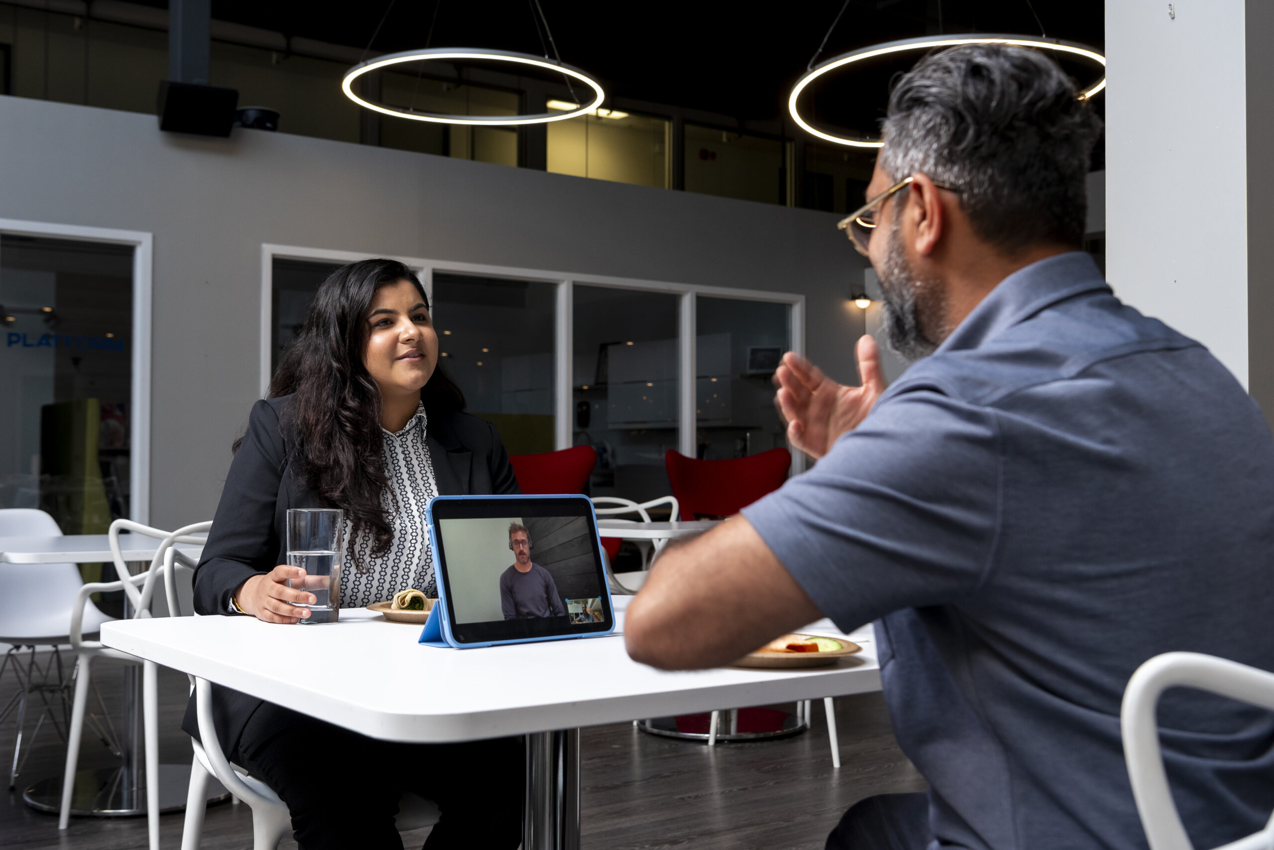 Two colleagues sit at a white cafeteria table having a conversation. The man signs while a tablet displays a Video Remote Interpreter on the table. The woman, wearing a black blazer, listens attentively with a glass of water and a snack in front of her. Circular lights hang from the ceiling in the modern space.